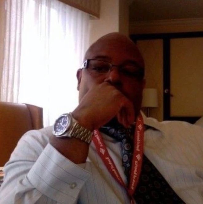 Person in glasses sitting indoors, wearing a tie and lanyard, resting chin on hand.